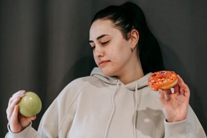 Pensive female wearing casual hoodie deciding between healthy green apple and sweet doughnut in studio