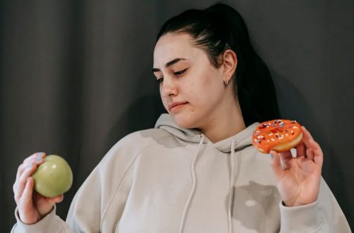 Pensive female wearing casual hoodie deciding between healthy green apple and sweet doughnut in studio